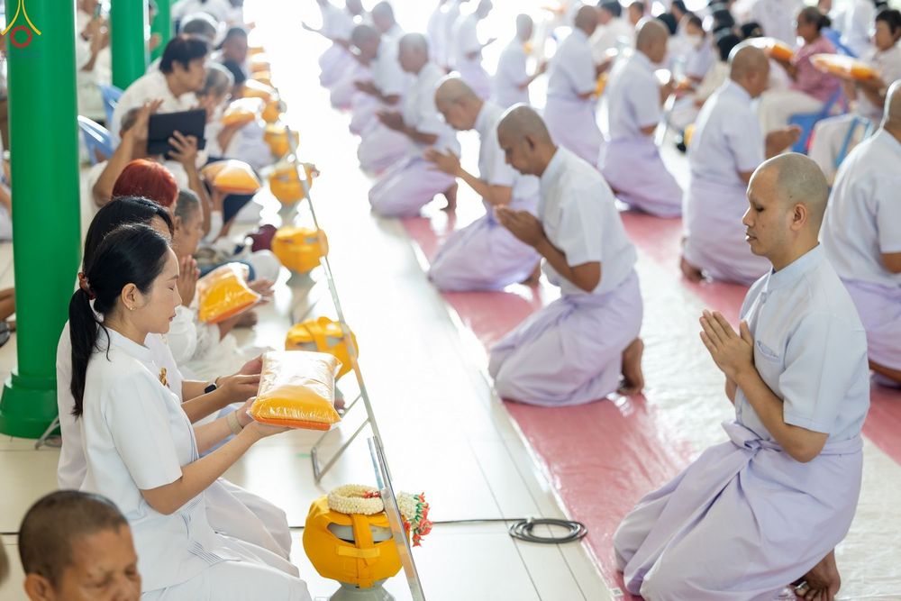 ผู้เข้าร่วมพิธีอุปสมบทหมู่ในชุดสีขาวกำลังนั่งพนมมือและประเคนผ้าไตรจีวรภายในโบสถ์