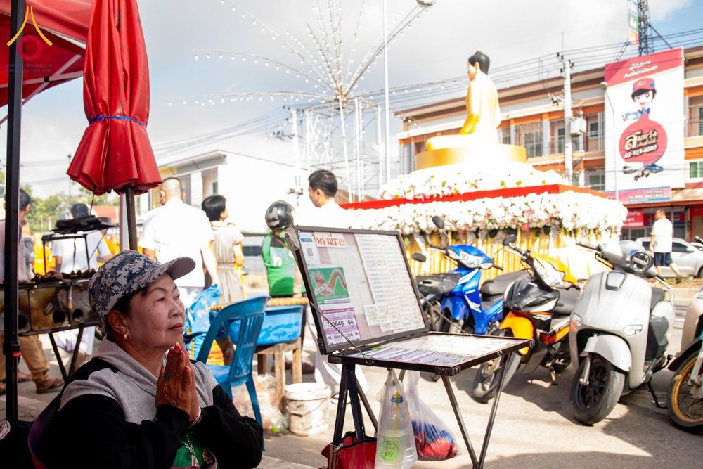 หญิงสูงวัยพนมมือไหว้พระพุทธรูปองค์สีทองที่ประดิษฐานอยู่บนฐานดอกไม้สีขาวท่ามกลางบรรยากาศงานพิธี