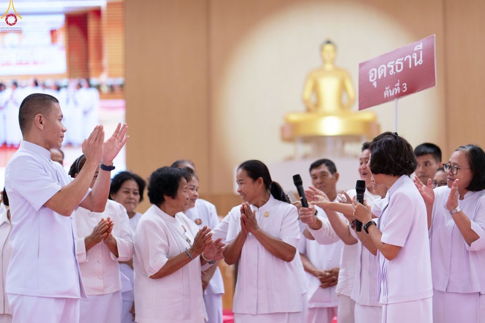 กลุ่มคนสวมชุดสีขาวกำลังยืนพนมมือและถือไมโครโฟนในงานสัมมนา โดยมีป้ายชื่อจังหวัดอุดรธานีและพระพุทธรูปเป็นฉากหลัง