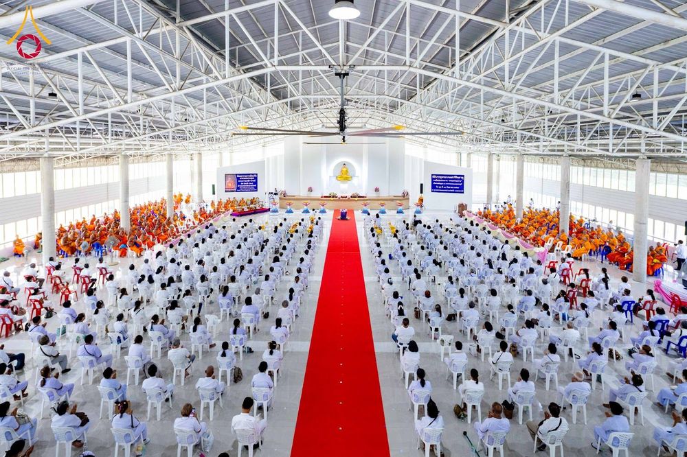 พุทธศาสนิกชนจำนวนมากสวมชุดขาวนั่งบนเก้าอี้ภายในอาคารโถงกว้างเพื่อร่วมพิธีทางศาสนา