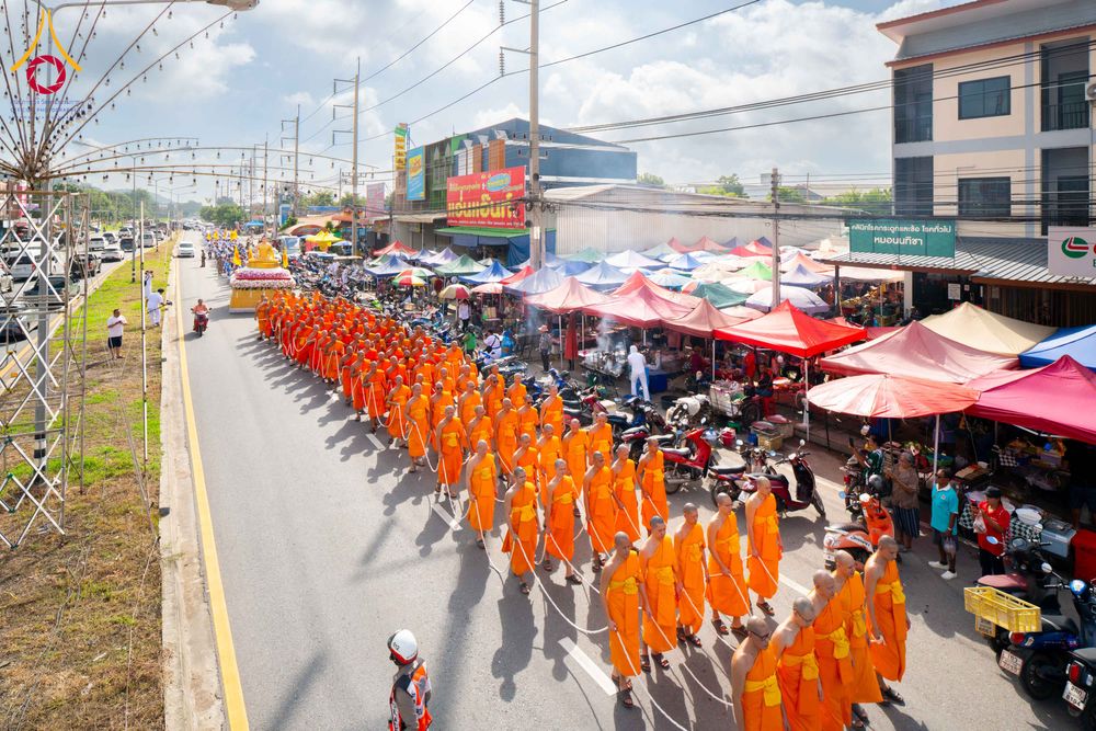 ขบวนพระภิกษุสงฆ์จำนวนมากเดินเรียงแถวบนถนนในวันที่มีแดดจัด โดยมีรถอัญเชิญพระประธานนำหน้าขบวนและมีร้านค้าตั้งอยู่ริมทาง