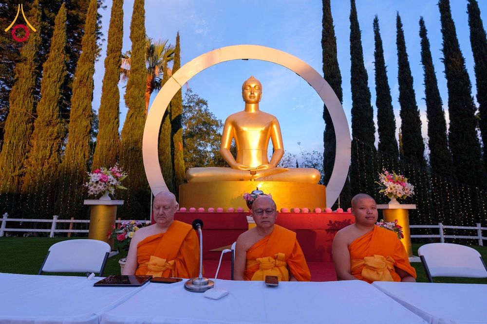 พระภิกษุสงฆ์สามรูปนั่งอยู่หน้าพระพุทธรูปสีทองขนาดใหญ่ในพื้นที่กลางแจ้ง