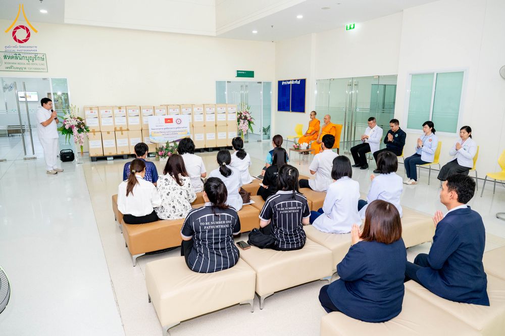 กลุ่มบุคคลและพระภิกษุสงฆ์นั่งร่วมในพิธีมอบยาและเวชภัณฑ์ภายในอาคารบุญรักษา