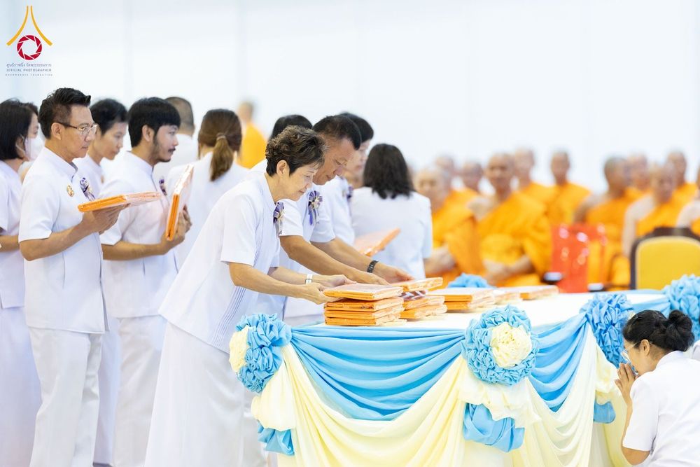 กลุ่มพุทธศาสนิกชนในชุดสีขาวกำลังวางผ้าไตรบนโต๊ะที่มีการตกแต่งด้วยผ้าสีฟ้าและสีเหลือง ในพิธีทางศาสนา