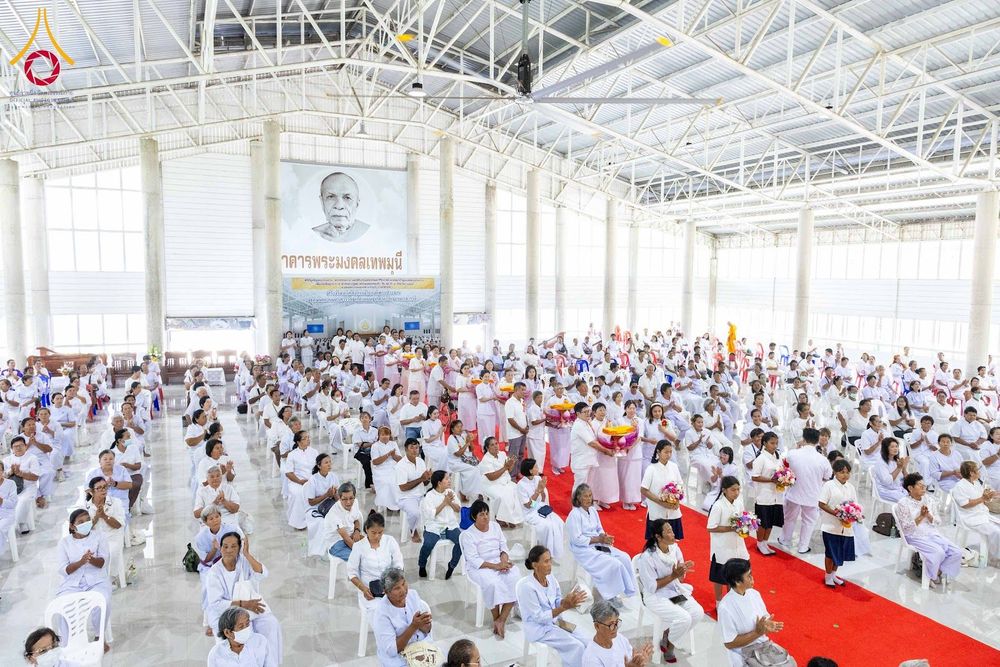 พุทธศาสนิกชนชุดขาวนั่งและยืนร่วมพิธีทางศาสนาภายในอาคารโถงกว้างที่มีพรมแดงปูพื้น