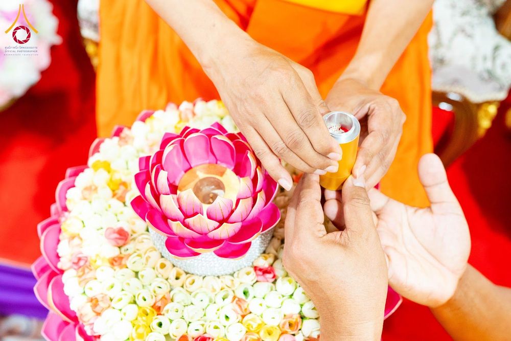 มือของพระภิกษุและฆราวาสกำลังประกอบพิธีบรรจุพระบรมสารีริกธาตุลงในภาชนะรูปดอกบัวตูมบนพานพุ่มดอกไม้