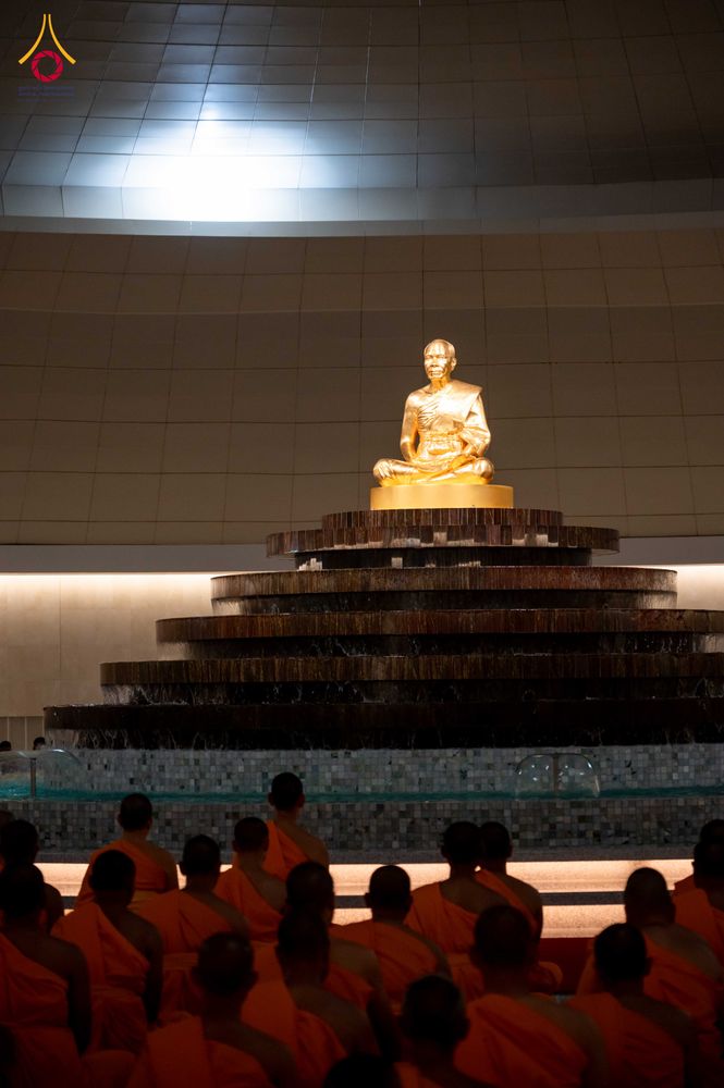 พระพุทธรูปสีทองประดิษฐานอยู่บนฐานเหนือสระน้ำ โดยมีกลุ่มผู้ปฏิบัติธรรมในชุดจีวรสีส้มนั่งอยู่ด้านหน้า