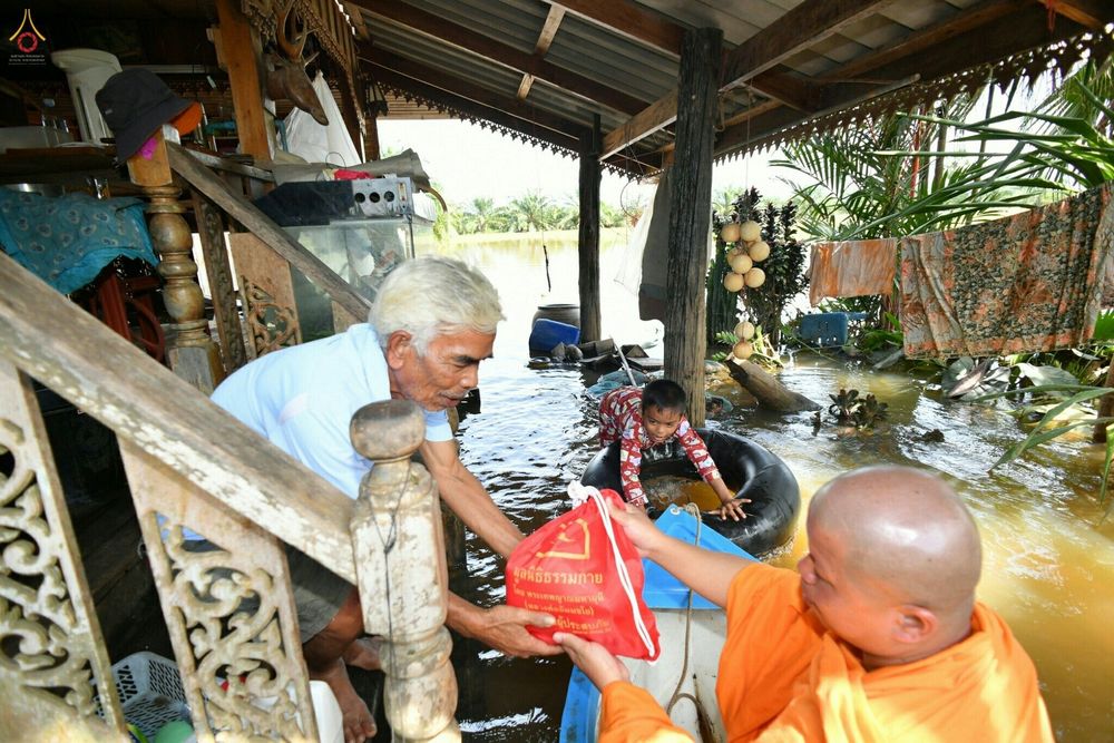 พระภิกษุในชุดจีวรสีส้มกำลังส่งถุงยังชีพสีแดงให้แก่ชายสูงวัยบนบ้านไม้ที่ถูกน้ำท่วมขัง โดยมีเด็กชายลอยคออยู่ในห่วงยางใกล้เรือ
