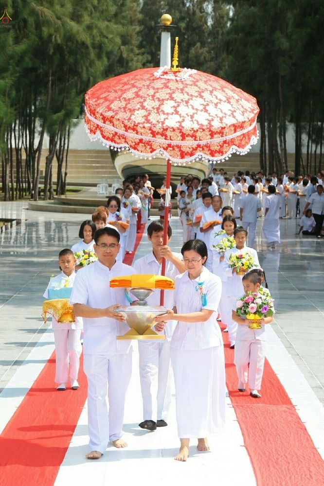กลุ่มพุทธศาสนิกชนในชุดขาวกำลังเดินในขบวนอัญเชิญผ้าไตรกฐินบนพรมแดง โดยมีผู้ถือร่มสีแดงนำขบวนในงานบุญ
