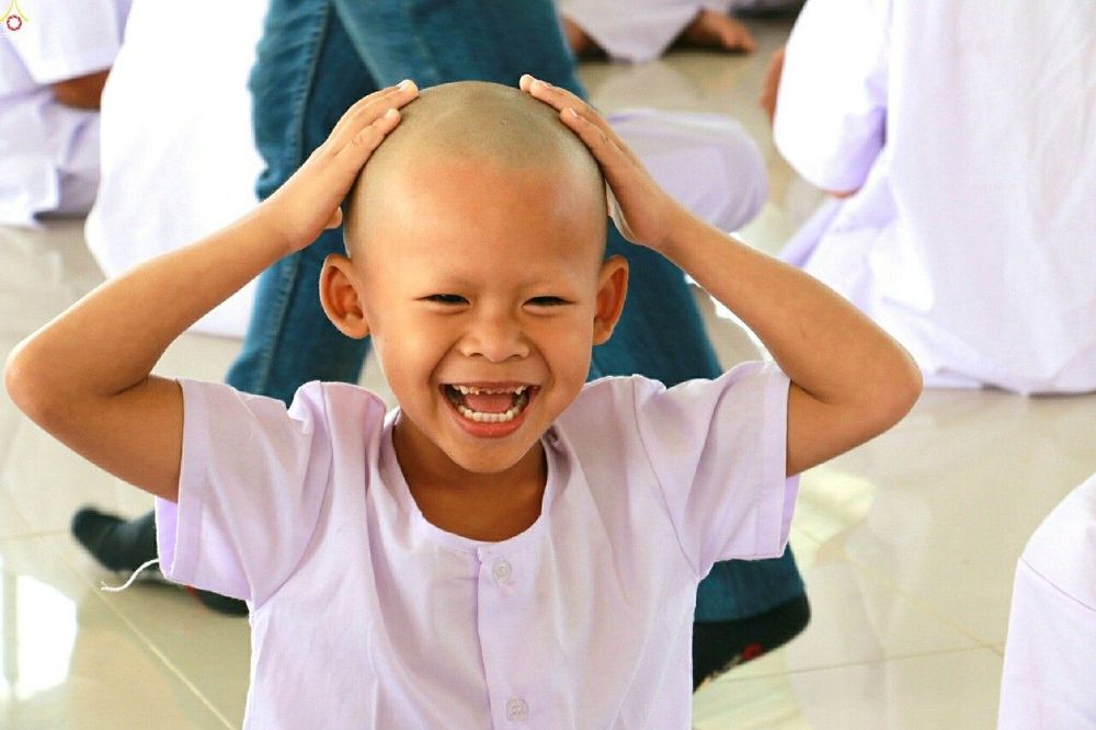 เด็กชายโกนศีรษะสวมชุดขาวกำลังยิ้มแย้มอย่างมีความสุขในพิธีปลงผมนาค