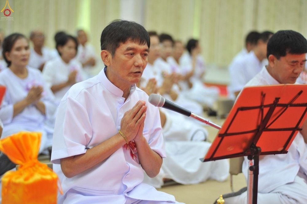 ชายไทยสวมชุดขาวพนมมือกล่าวคำบูชาผ่านไมโครโฟนในพิธีทอดกฐินสามัคคี โดยมีพุทธศาสนิกชนชุดขาวนั่งล้อมรอบ