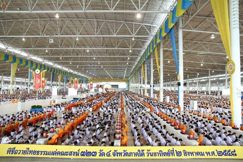 พุทธศาสนิกชนจำนวนมากนั่งเรียงแถวประกอบพิธีถวายไทยธรรมแด่คณะสงฆ์ภายในอาคารขนาดใหญ่