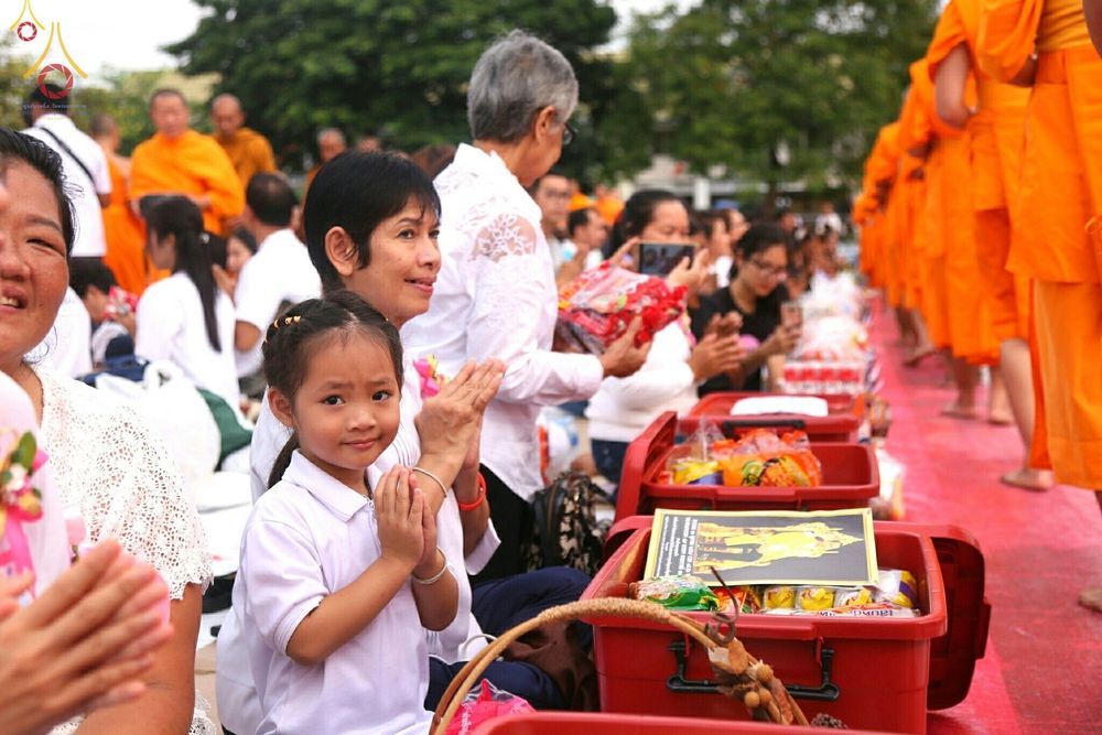 เด็กหญิงและผู้ใหญ่ในชุดสีขาวกำลังพนมมือรอใส่บาตรพระสงฆ์จำนวนมากที่เดินเรียงแถวมาในพิธีตักบาตร