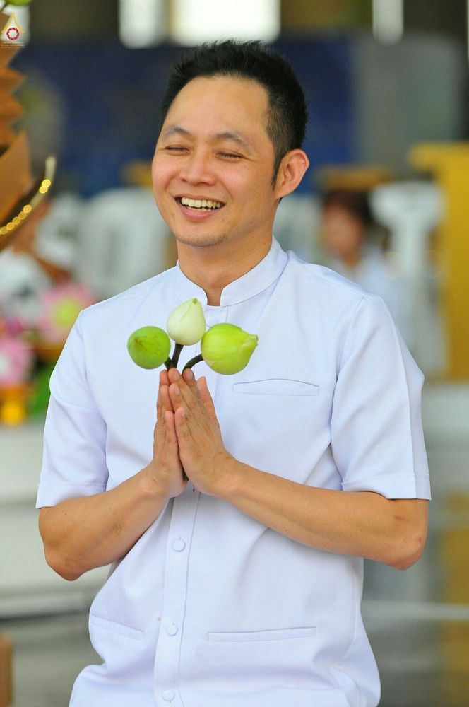 ชายไทยสวมชุดขาวพนมมือถือดอกบัวด้วยใบหน้ายิ้มแย้มในพิธีกรรมทางศาสนา