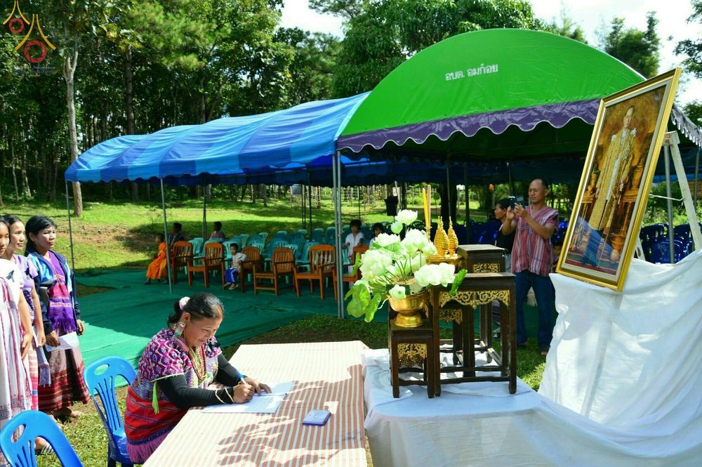 หญิงชาวไทยภูเขาแต่งกายชุดพื้นเมืองกำลังลงนามในสมุดบันทึกหน้าโต๊ะหมู่บูชาที่มีพระบรมฉายาลักษณ์ตั้งอยู่ ในงานพิธีทอดกฐินสามัคคี วัดทุ่งเจริญธรรม จังหวัดเชียงใหม่