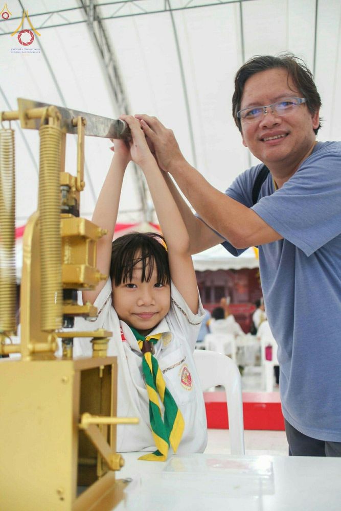 เด็กหญิงในชุดลูกเสือและชายสวมแว่นตากำลังช่วยกันใช้เครื่องกดพิมพ์พระของขวัญ