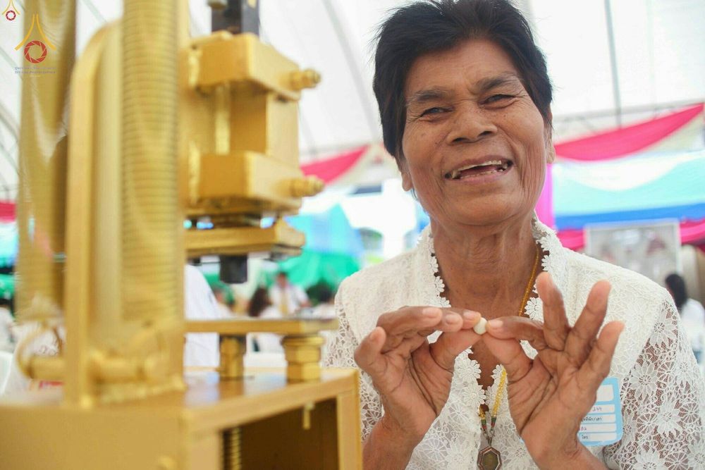 หญิงสูงวัยสวมชุดสีขาวกำลังยิ้มแย้มถือพระของขวัญขนาดเล็กที่เพิ่งพิมพ์เสร็จจากเครื่องกดพระสีทอง