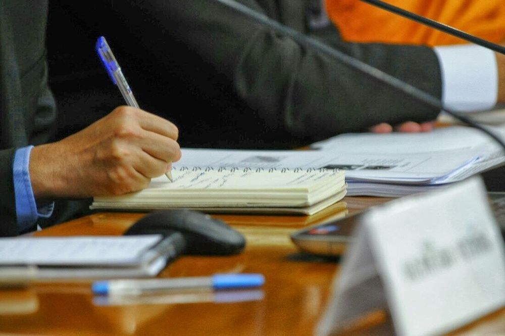 มือของผู้เข้าร่วมสัมมนาที่กำลังใช้ปากกาจดบันทึกข้อมูลลงในสมุดโน้ตบนโต๊ะประชุมที่มีเอกสารและไมโครโฟนวางอยู่