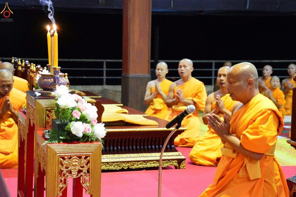 พระสงฆ์กำลังนั่งพนมมือสวดมนต์ในพิธีหล่อเทียนพรรษาโดยมีโต๊ะหมู่บูชาและเทียนที่จุดไฟอยู่ด้านหน้า