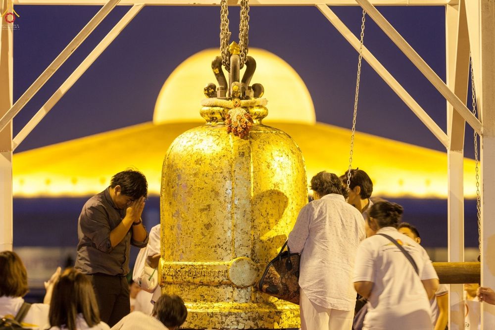 พุทธศาสนิกชนกำลังปิดแผ่นทองบนระฆังธรรมขนาดใหญ่โดยมีมหาเจดีย์สีทองเป็นฉากหลัง