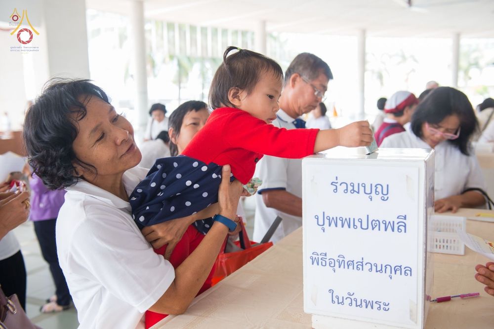 เด็กหญิงสวมเสื้อสีแดงกำลังหย่อนเงินลงในกล่องรับบริจาคพิธีบุพเปตพลีโดยมีผู้ใหญ่อุ้มอยู่
