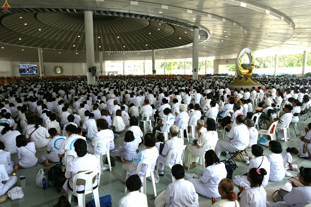 พุทธศาสนิกชนสวมชุดขาวนั่งเก้าอี้ร่วมพิธีทำบุญภายในอาคารทรงกลมขนาดใหญ่