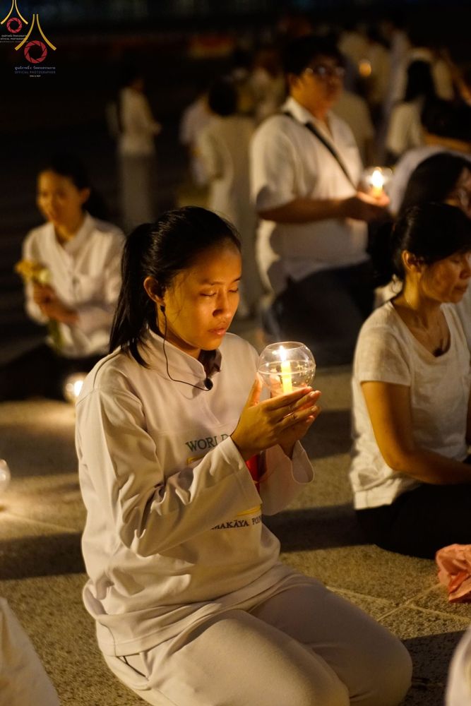 หญิงสาวสวมชุดขาวถือโคมประทีปในมือขณะนั่งสมาธิร่วมกับพุทธศาสนิกชนท่านอื่นในพิธีสวดมนต์ยามค่ำคืน
