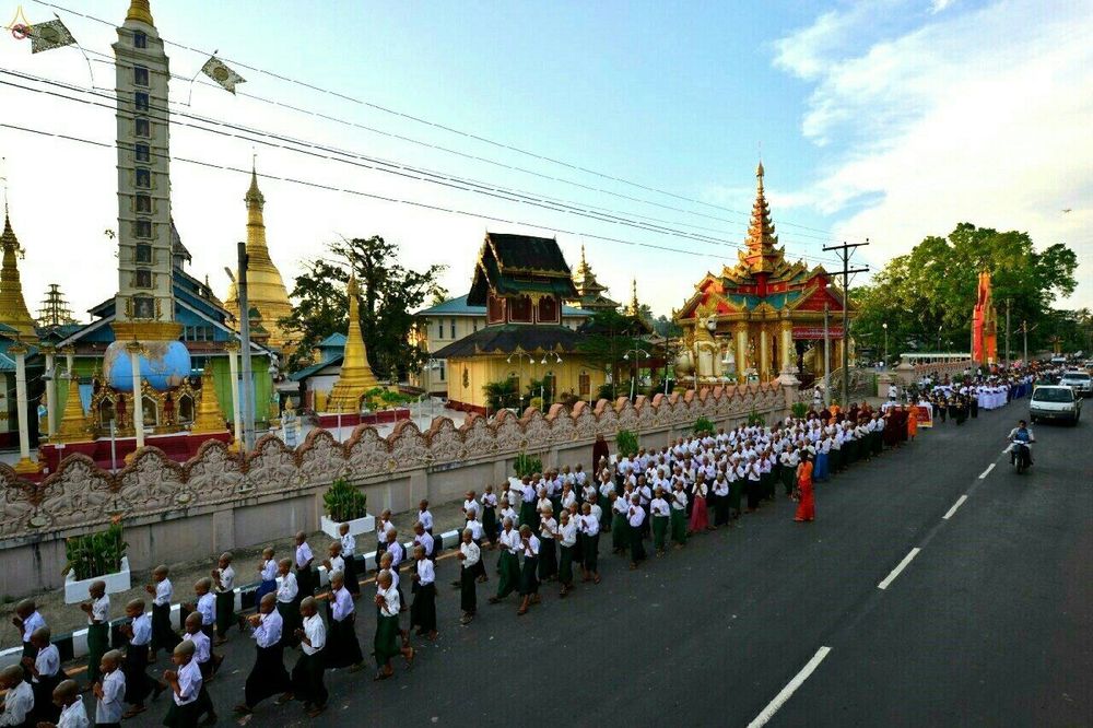 ขบวนนาคและผู้เข้าร่วมพิธีบรรพชาสามเณรเดินแถวบนถนนในเมืองทวายโดยมีฉากหลังเป็นวัดพม่า