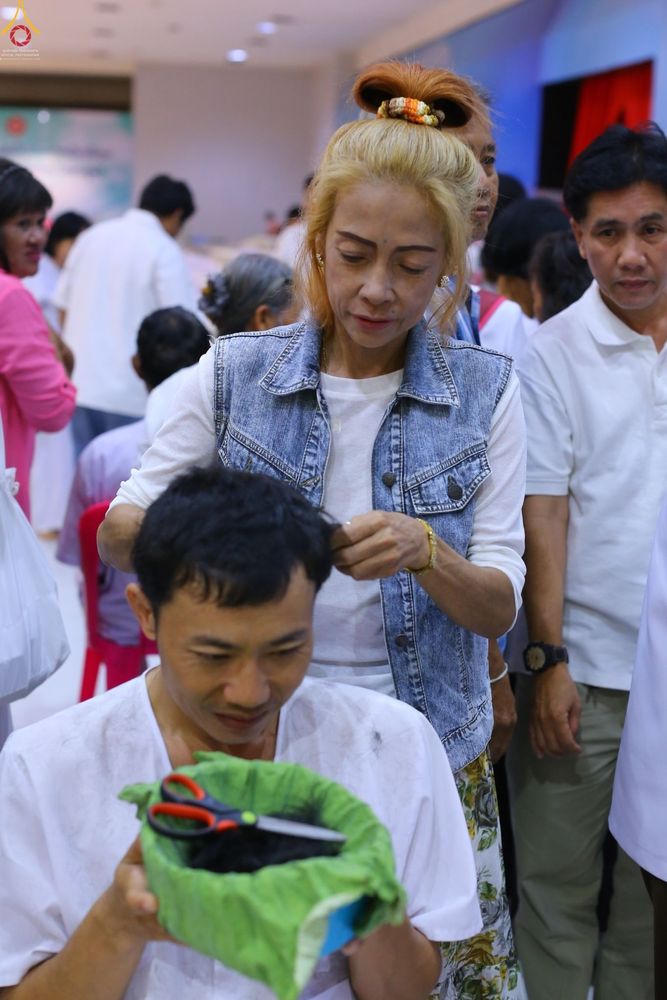 หญิงวัยกลางคนกำลังใช้กรรไกรตัดปอยผมให้แก่ชายหนุ่มที่สวมชุดขาวเตรียมอุปสมบท โดยมีกรรไกรวางอยู่บนพานรองรับเส้นผม