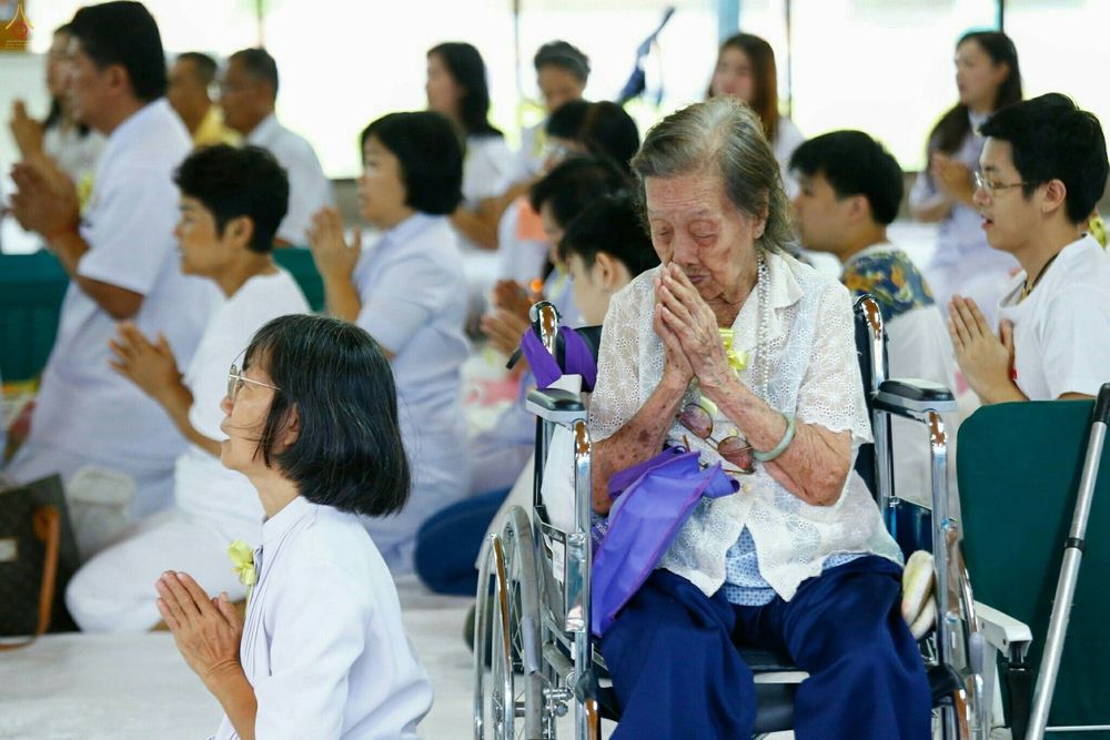 กลุ่มพุทธศาสนิกชนแต่งกายชุดขาวกำลังพนมมือทำบุญ โดยมีหญิงชรานั่งบนรถเข็นเป็นจุดเด่นในภาพ