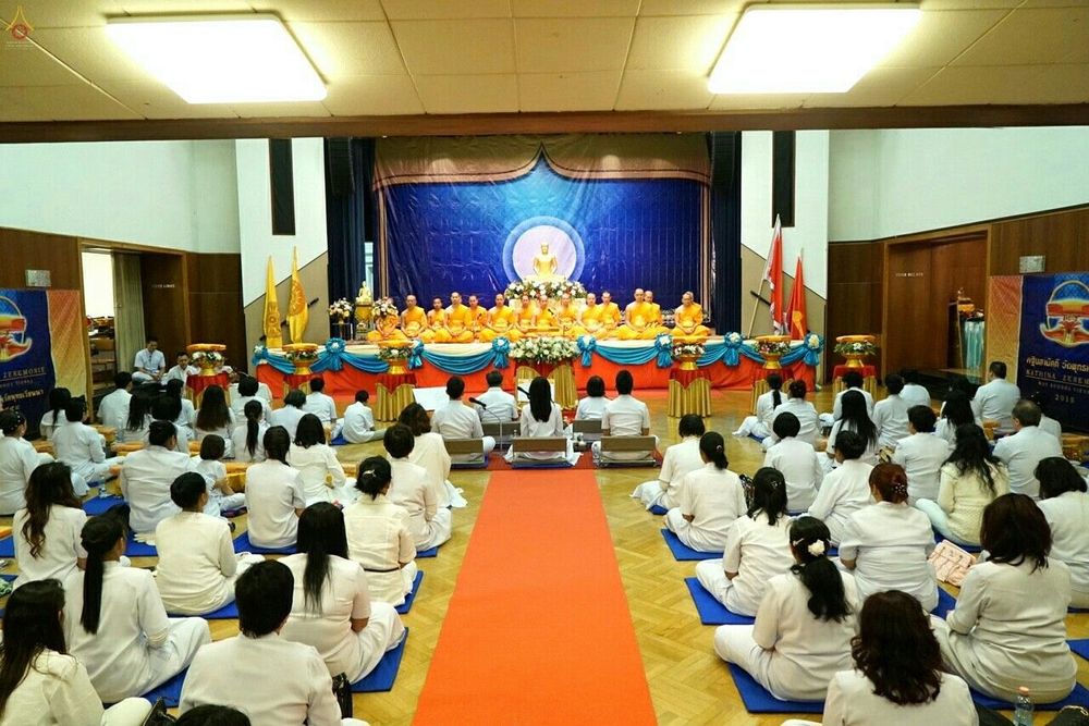 พุทธศาสนิกชนสวมชุดขาวนั่งพนมมือร่วมพิธีทอดกฐินภายในหอประชุม โดยมีคณะสงฆ์นั่งบนเวทีด้านหน้า