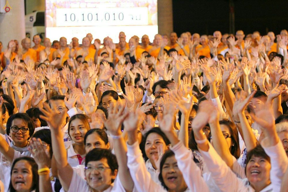 กลุ่มพุทธศาสนิกชนสวมชุดขาวชูมือขึ้นพร้อมกันในพิธีสวดธัมมจักกัปปวัตนสูตร โดยมีป้ายแสดงยอดการสวด ๑๐,๑๐๑,๐๑๐ จบเป็นฉากหลัง