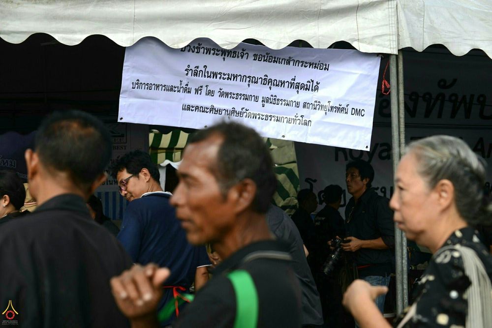 ประชาชนเดินผ่านจุดบริการอาหารและน้ำดื่มฟรีที่มีป้ายข้อความรำลึกในพระมหากรุณาธิคุณ