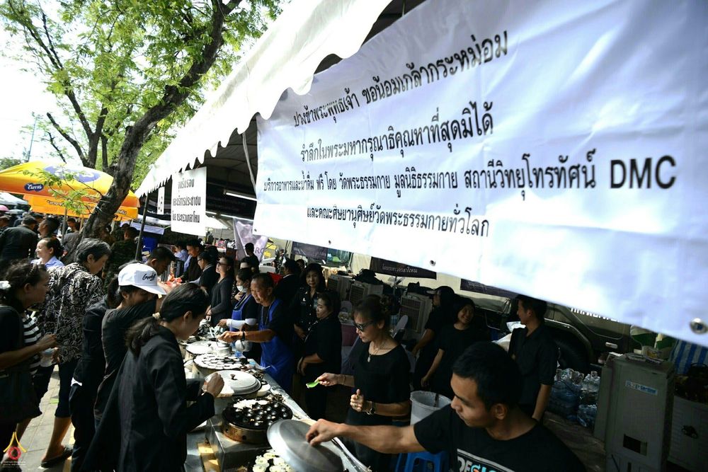 กลุ่มจิตอาสากำลังปรุงอาหารและแจกจ่ายน้ำดื่มให้แก่ประชาชนที่มาเข้าแถวรับบริการภายใต้เต็นท์สีขาว
