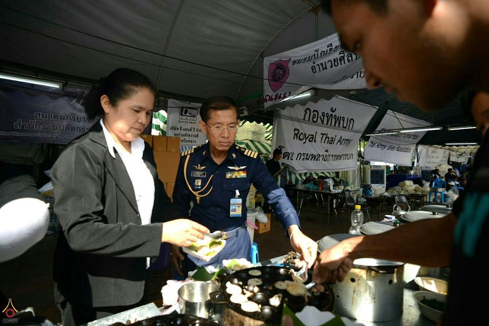 เจ้าหน้าที่ทหารและหญิงในชุดสุภาพกำลังช่วยกันทำขนมครกเพื่อแจกจ่ายให้กับประชาชนที่มาสักการะพระบรมศพ