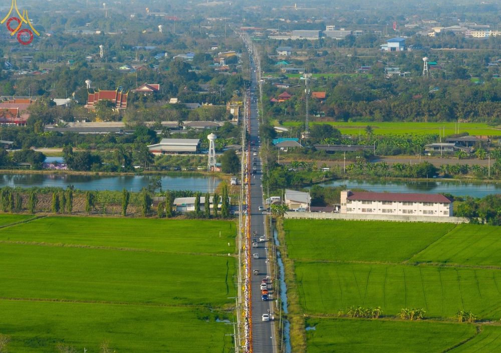 ขบวนพระภิกษุสงฆ์เดินธุดงค์เป็นแถวยาวบนถนนท่ามกลางทุ่งนาสีเขียวขจีในมุมมองจากที่สูง