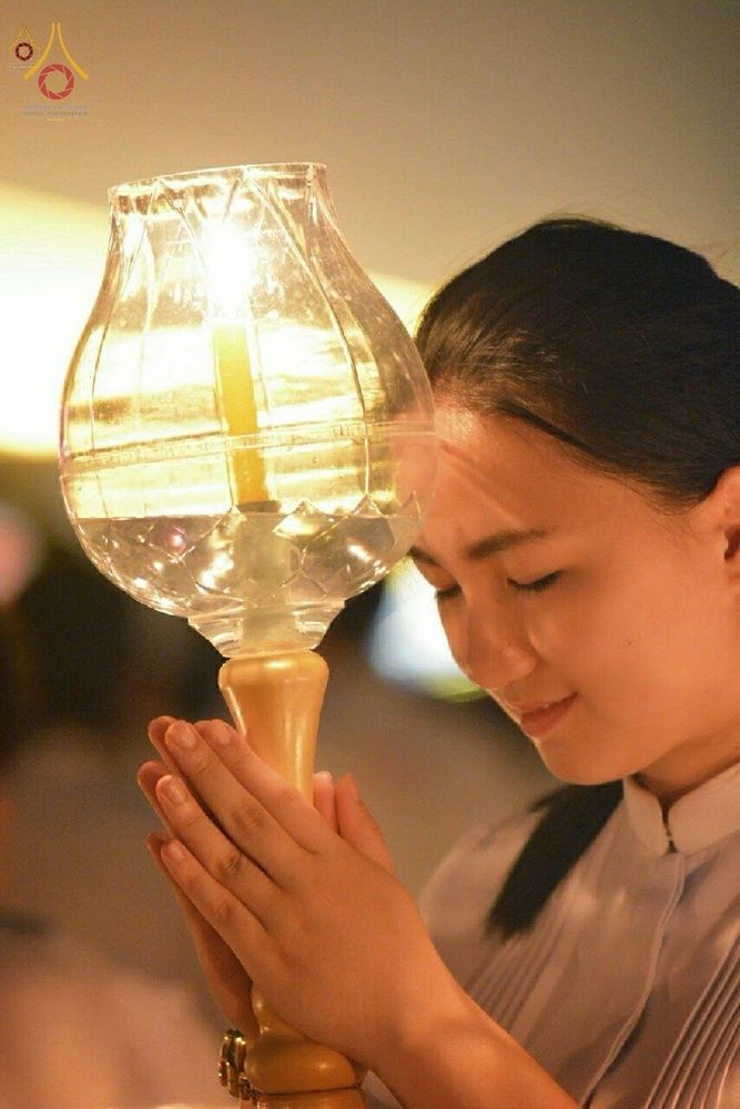 หญิงสาวสวมชุดสีขาวประนมมือถือโคมประทีปแก้วที่มีแสงเทียนสว่างไสวด้วยความศรัทธา