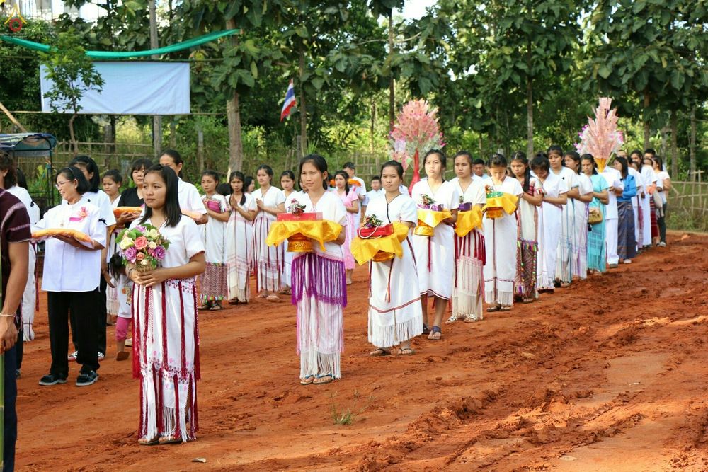 กลุ่มหญิงสาวในชุดพื้นเมืองสีขาวถือพานพุ่มและดอกไม้เดินเรียงแถวในพิธีทอดกฐิน