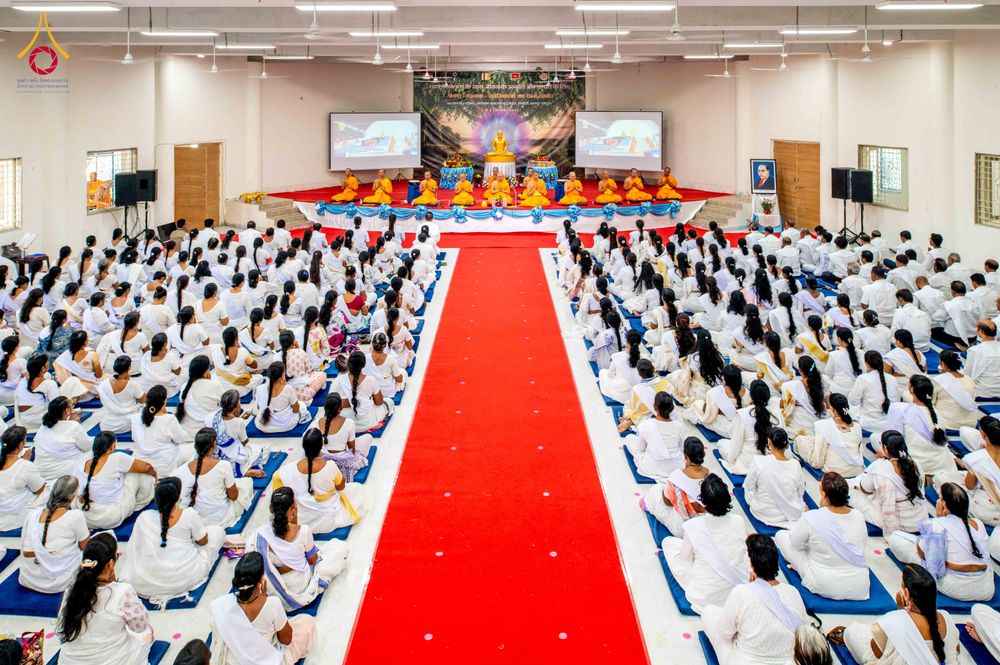 กลุ่มผู้เข้าร่วมอบรมในชุดสีขาวนั่งเรียงรายในห้องโถงกว้างโดยมีพรมสีแดงทอดยาวไปสู่เวทีที่มีคณะสงฆ์นั่งอยู่