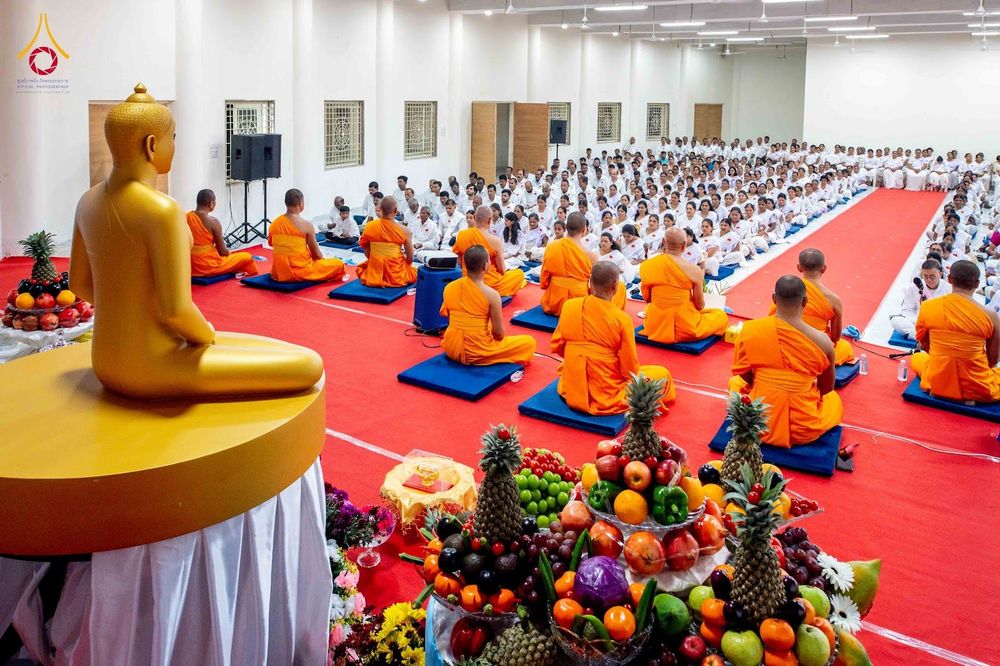 พระภิกษุสงฆ์และผู้เข้าร่วมโครงการในชุดสีขาวนั่งปฏิบัติธรรมภายในหอประชุมที่มีพรมสีแดงและพระพุทธรูปสีทอง