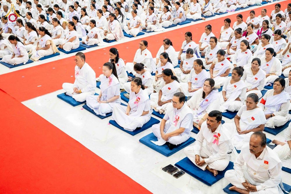 กลุ่มคนในชุดสีขาวนั่งสมาธิบนเบาะรองนั่งในอาคารที่มีพรมสีแดง