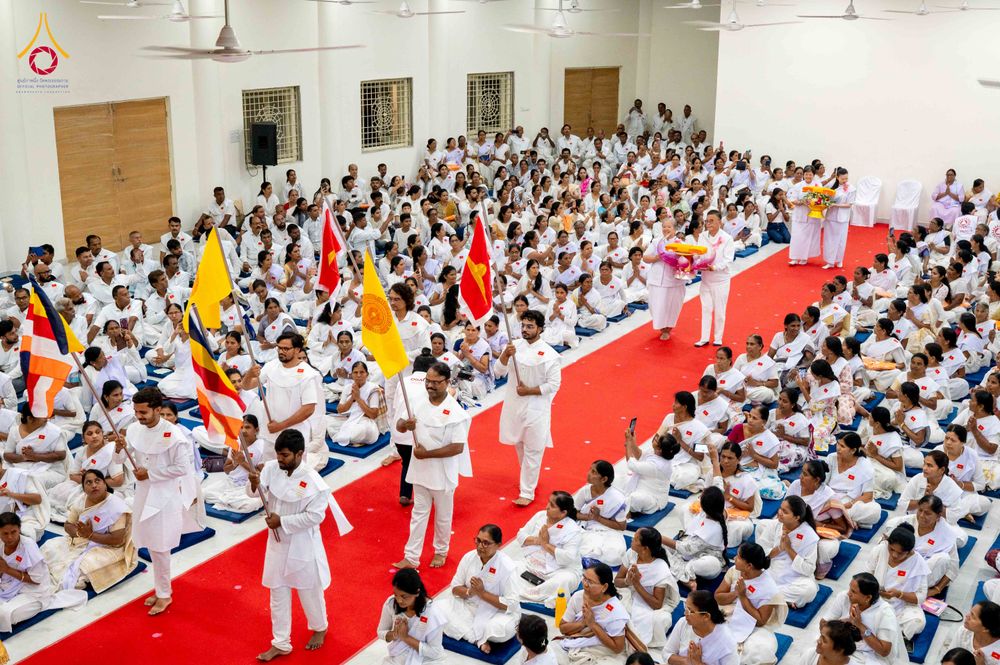 กลุ่มคนสวมชุดสีขาวนั่งเรียงรายในอาคาร มีขบวนถือธงและพานดอกไม้เดินบนพรมแดงในพิธีปิดโครงการอบรม