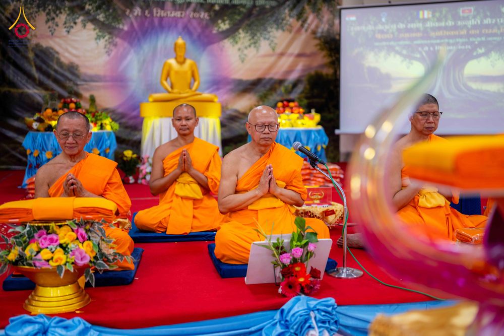 พระภิกษุสงฆ์สามรูปนั่งพนมมือในพิธีทางศาสนาพุทธ เบื้องหลังเป็นพระพุทธรูปและฉากหลังภาพต้นโพธิ์