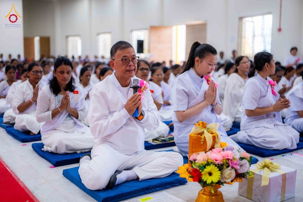 กลุ่มคนในชุดสีขาวนั่งพนมมือปฏิบัติธรรมภายในอาคาร โดยมีชายสวมแว่นตาถือไมโครโฟนเป็นผู้นำกล่าว