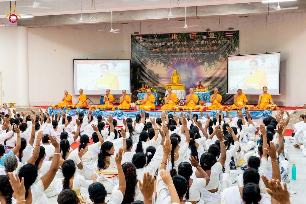 พระภิกษุสงฆ์นั่งบนเวทีหน้าป้ายโครงการและผู้เข้าร่วมอบรมในชุดสีขาวจำนวนมากนั่งหันหลังให้กล้องภายในหอประชุม