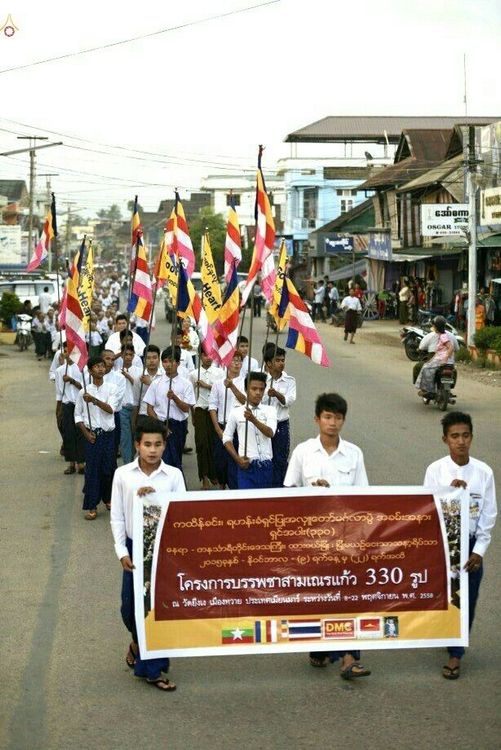 กลุ่มเยาวชนชายถือป้ายโครงการบรรพชาสามเณรแก้วและธงธรรมจักรเดินขบวนแห่นาคบนถนนในเมืองทวาย