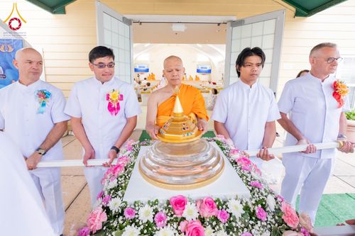 พระภิกษุสงฆ์และกลุ่มชายในชุดขาวกำลังช่วยกันประคองบุษบกที่บรรจุพระบรมสารีริกธาตุเพื่อนำเข้าสู่ภายในอาคาร