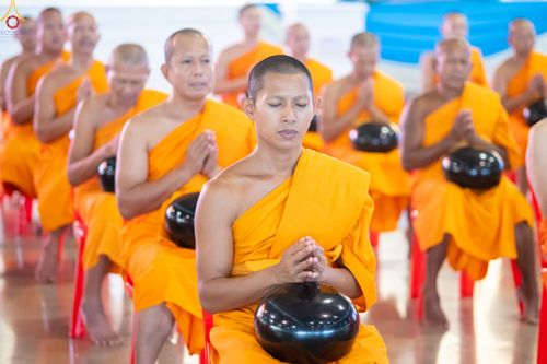 พระภิกษุสงฆ์หลายรูปสวมจีวรสีเหลืองนั่งพนมมือถือบาตรสีดำในพิธีทางศาสนา