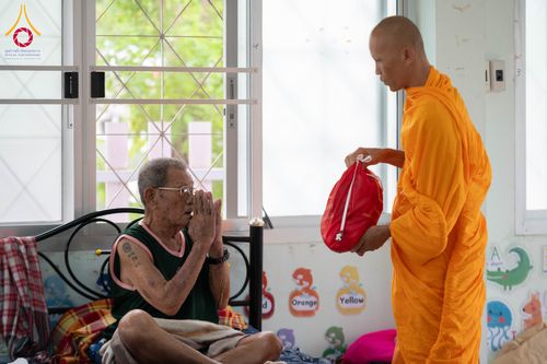 พระภิกษุส่งมอบถุงยังชีพสีแดงให้ผู้สูงอายุที่กำลังพนมมือรับบนเตียงนอน