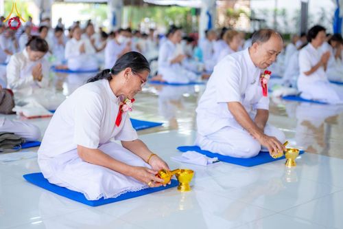 กลุ่มผู้ปฏิบัติธรรมในชุดสีขาวกำลังประกอบพิธีกรวดน้ำด้วยกรวยน้ำสีทองภายในสถานที่ปฏิบัติธรรม